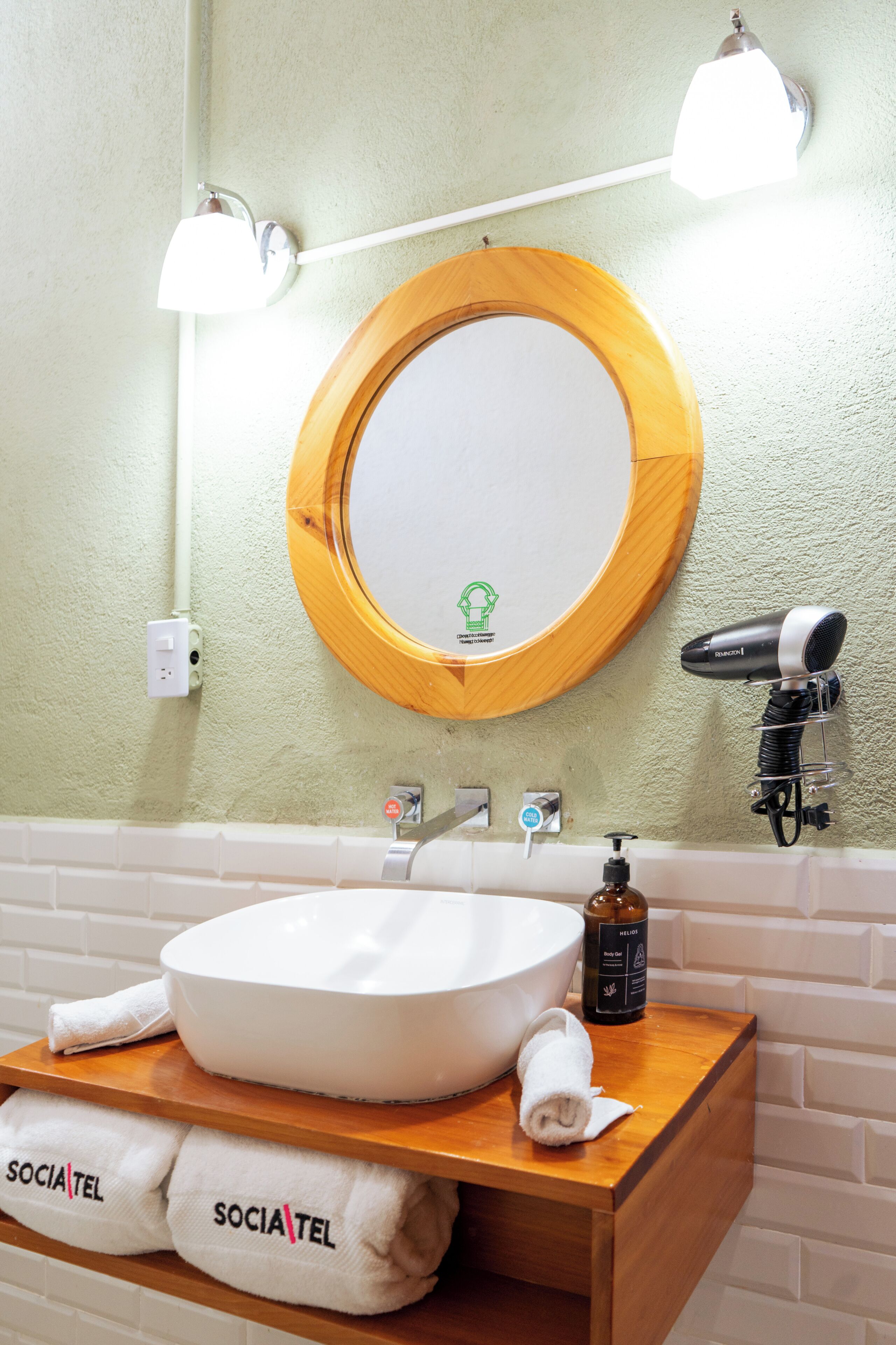 A bathroom with a round mirror, a white sink, a hairdryer, and towels labeled 'SOCIA TEL'.