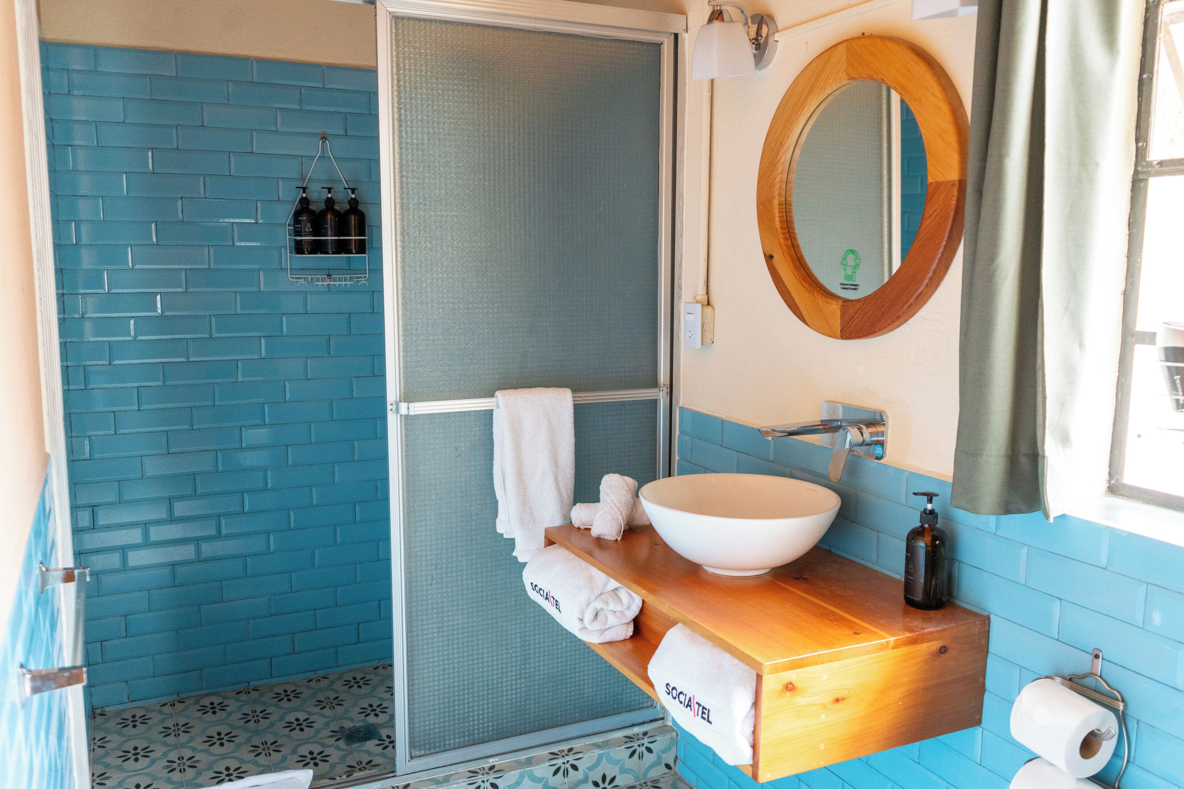 Une salle de bain avec un mur recouvert de carreaux bleus, un lavabo blanc, un meuble-lavabo en bois, une douche avec une porte en verre et un miroir avec un cadre en bois.