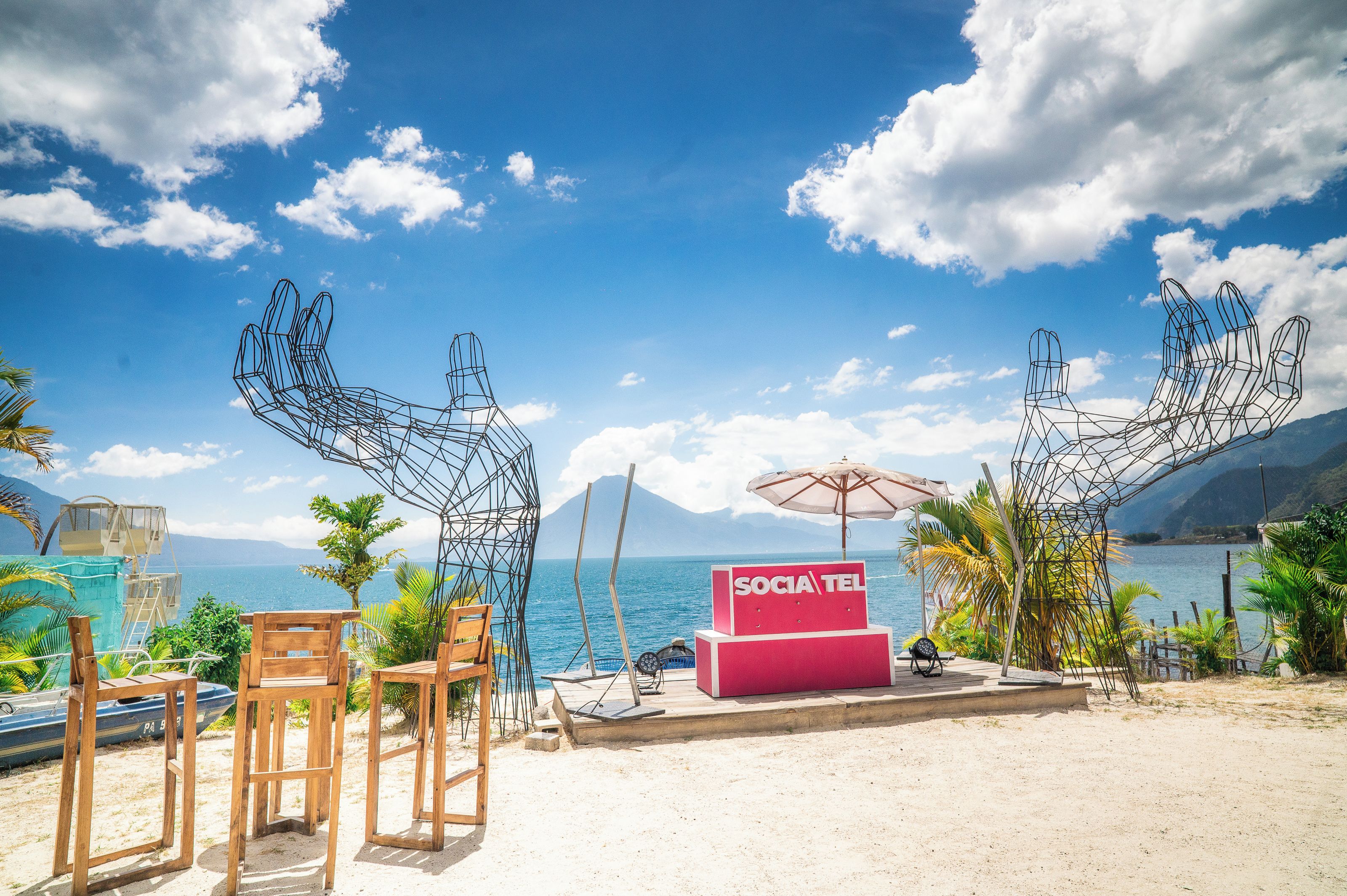 A beachside setting with a red 'SOCIA TEL' sign, wooden chairs, and a metal sculpture.