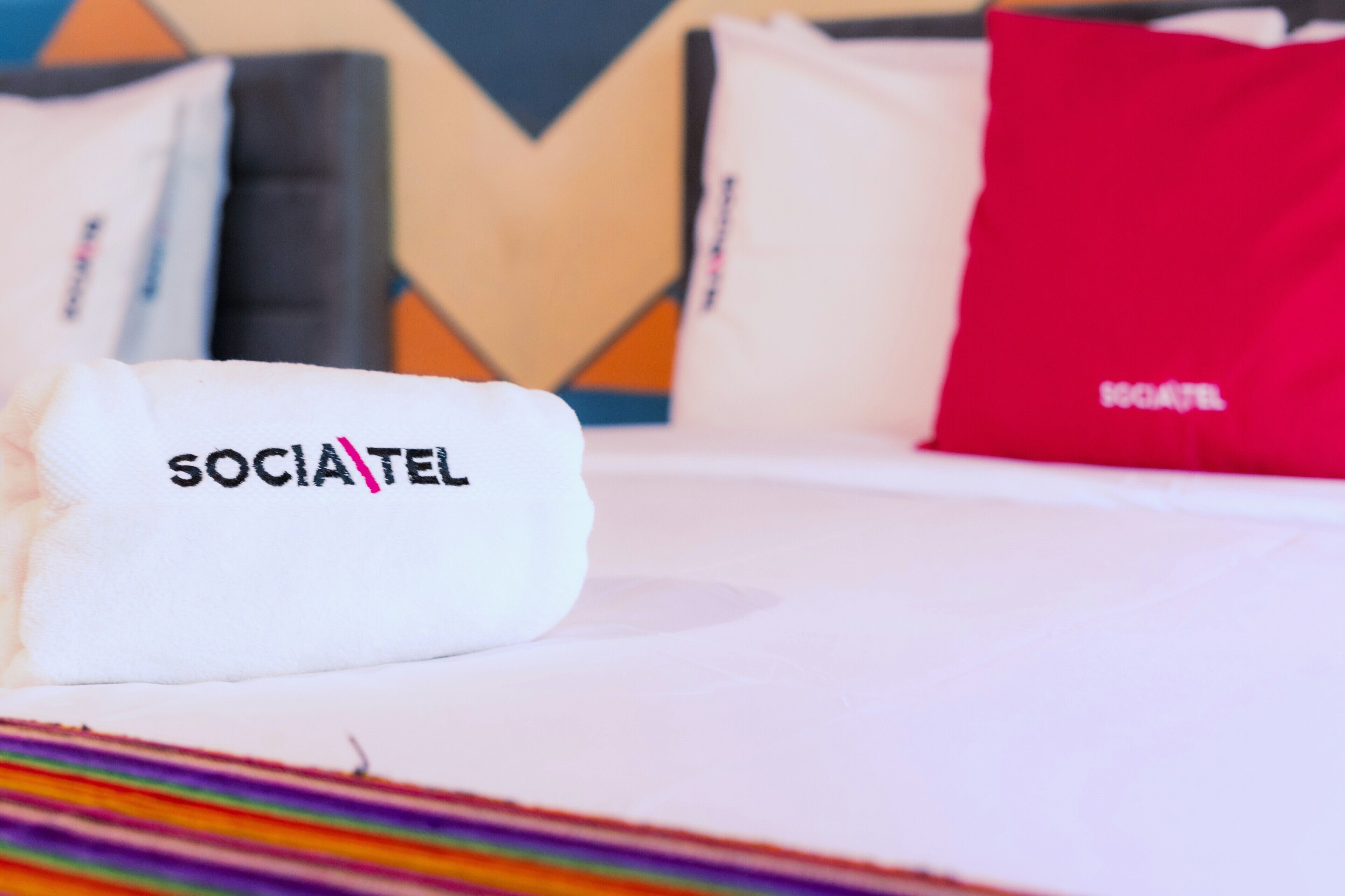 Una cama de hotel con una almohada blanca que dice 'SOCIATEL' y una almohada roja al fondo.