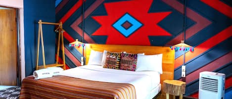 A hotel room with a bed, a wooden headboard, a small wooden stool, a wall-mounted air conditioner, and a patterned rug.