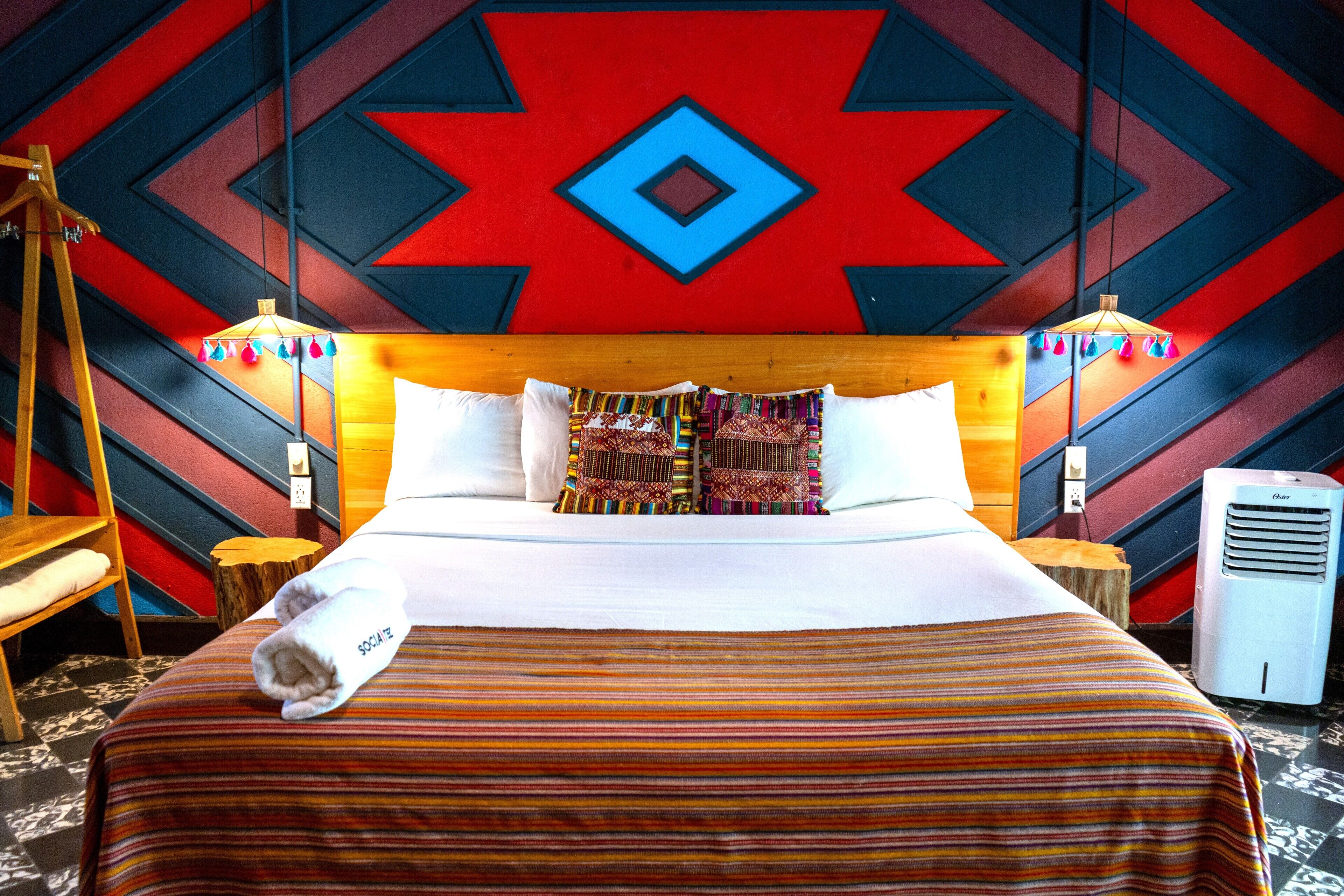 Ein Hotelzimmer mit einem großen Bett, einem Holzkopfteil, gemusterten Kissen, einem Handtuch auf dem Bett, zwei Nachttischen, einer Lampe und einer Wand mit geometrischem Muster.
