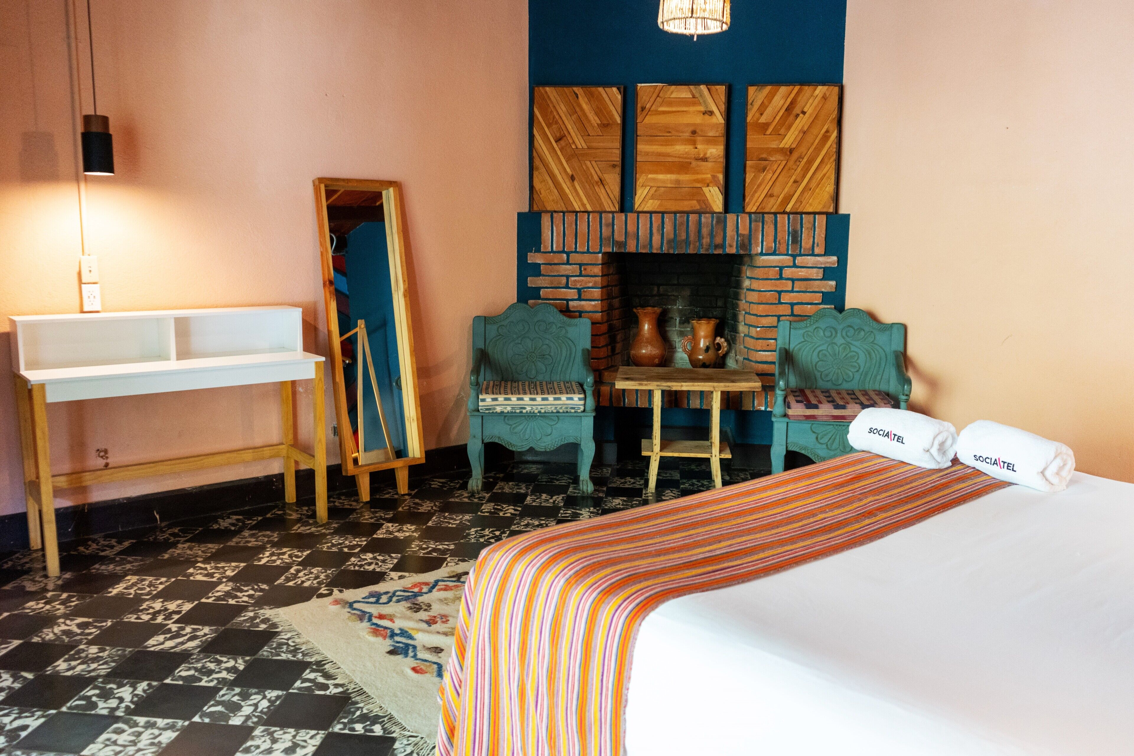 A hotel room with a bed, a small table with a vase, two chairs, a mirror, and a wall-mounted fireplace.
