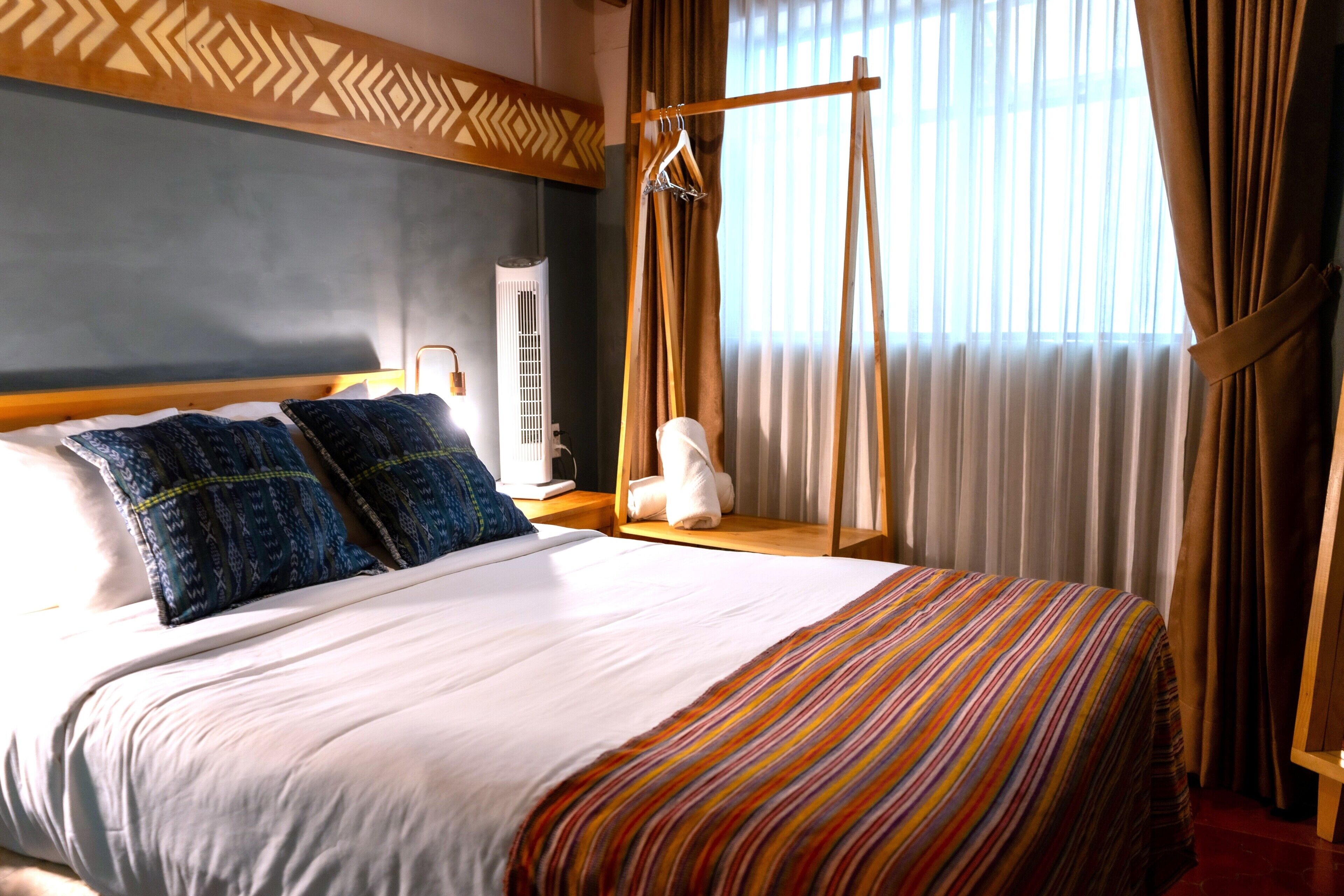 A hotel room with a bed, pillows, a bedside table, a lamp, a hanging garment rack, and a window with curtains.
