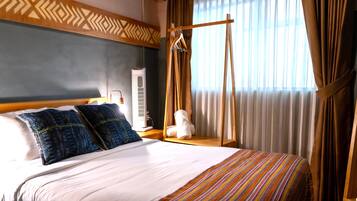 A hotel room with a bed, pillows, a bedside table, a lamp, a hanging garment rack, and a window with curtains.