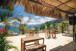 Un área de estar frente a la playa con mesas y sillas de madera, techo de paja y vista al mar y las montañas.