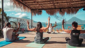 A group of people practicing yoga on a wooden deck with a thatched roof, overlooking a body of water and a mountain. - Socialtel Atitlan (Panajachel)