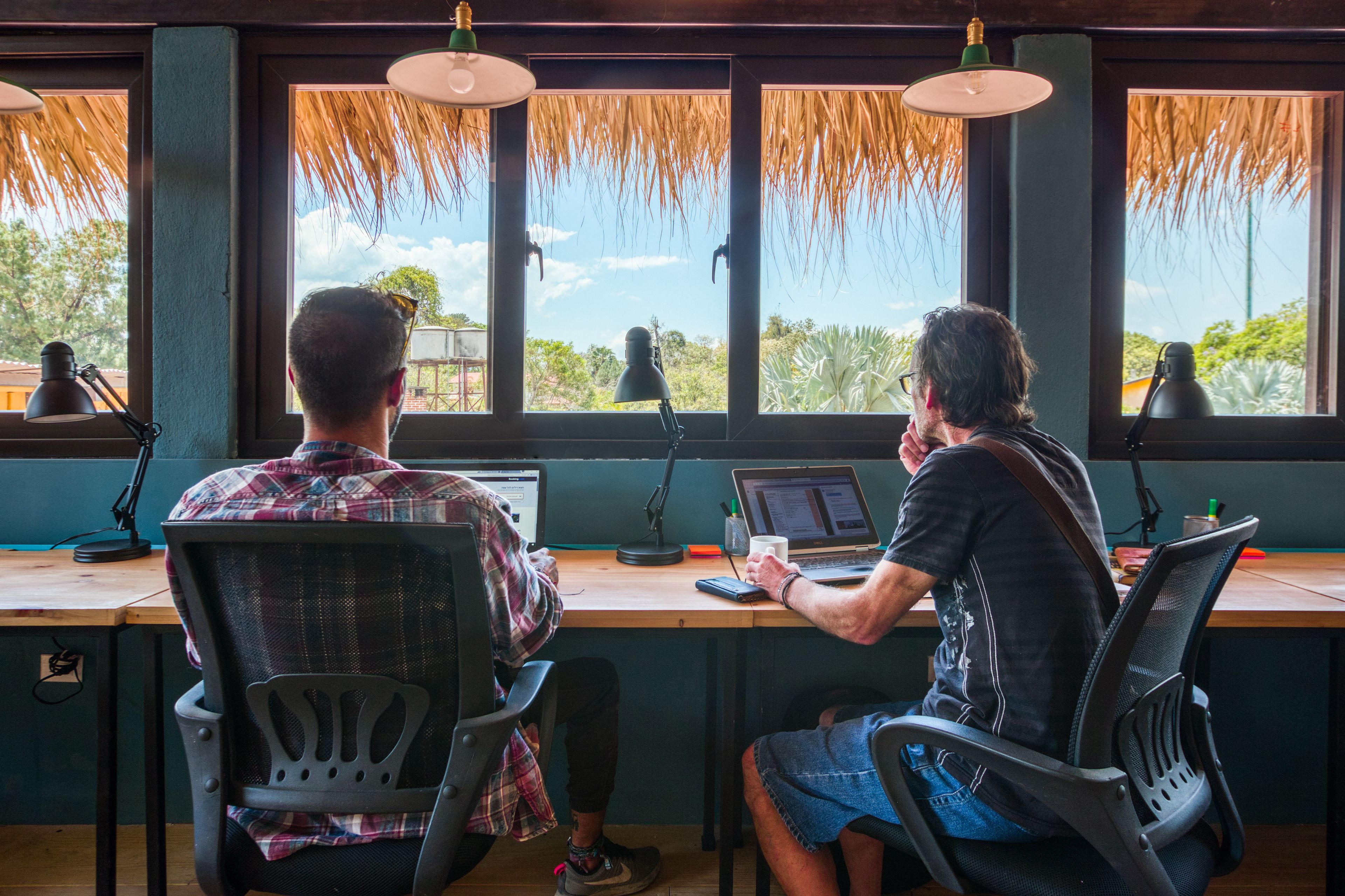 Zwei Personen arbeiten an Schreibtischen mit Computern, großen Fenstern und hängenden Lampen.
