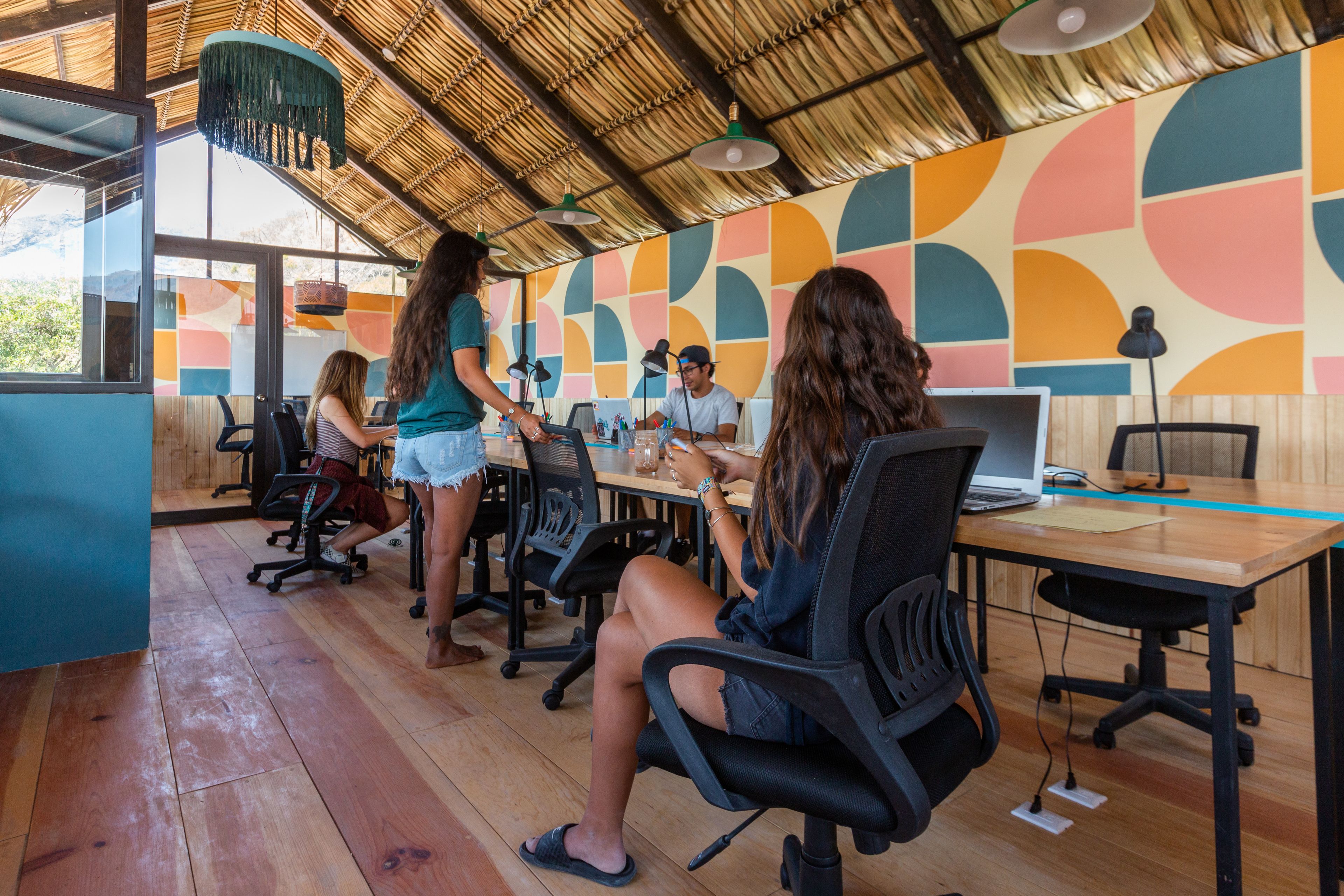 Un espace de travail partagé moderne, doté de planchers en bois, de chaises de bureau noires et d’un mur décoré de motifs géométriques colorés.