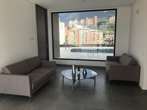 A modern living room with two sofas and a glass coffee table, large windows offering a view of high-rise buildings and a pool. - Hotel Class Suites By Colclick (Medellín)
