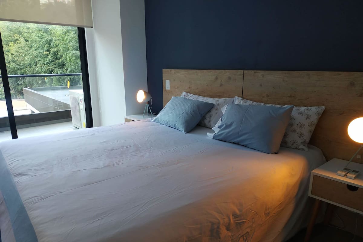 A bedroom with a large bed, two bedside lamps, a wooden headboard, and a view of greenery outside.
