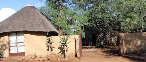 A thatched-roof house with a gated entrance, surrounded by trees and a dirt path.