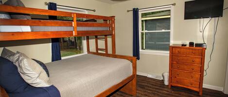 A room with a bunk bed, a dresser, a television, and a window with blinds.