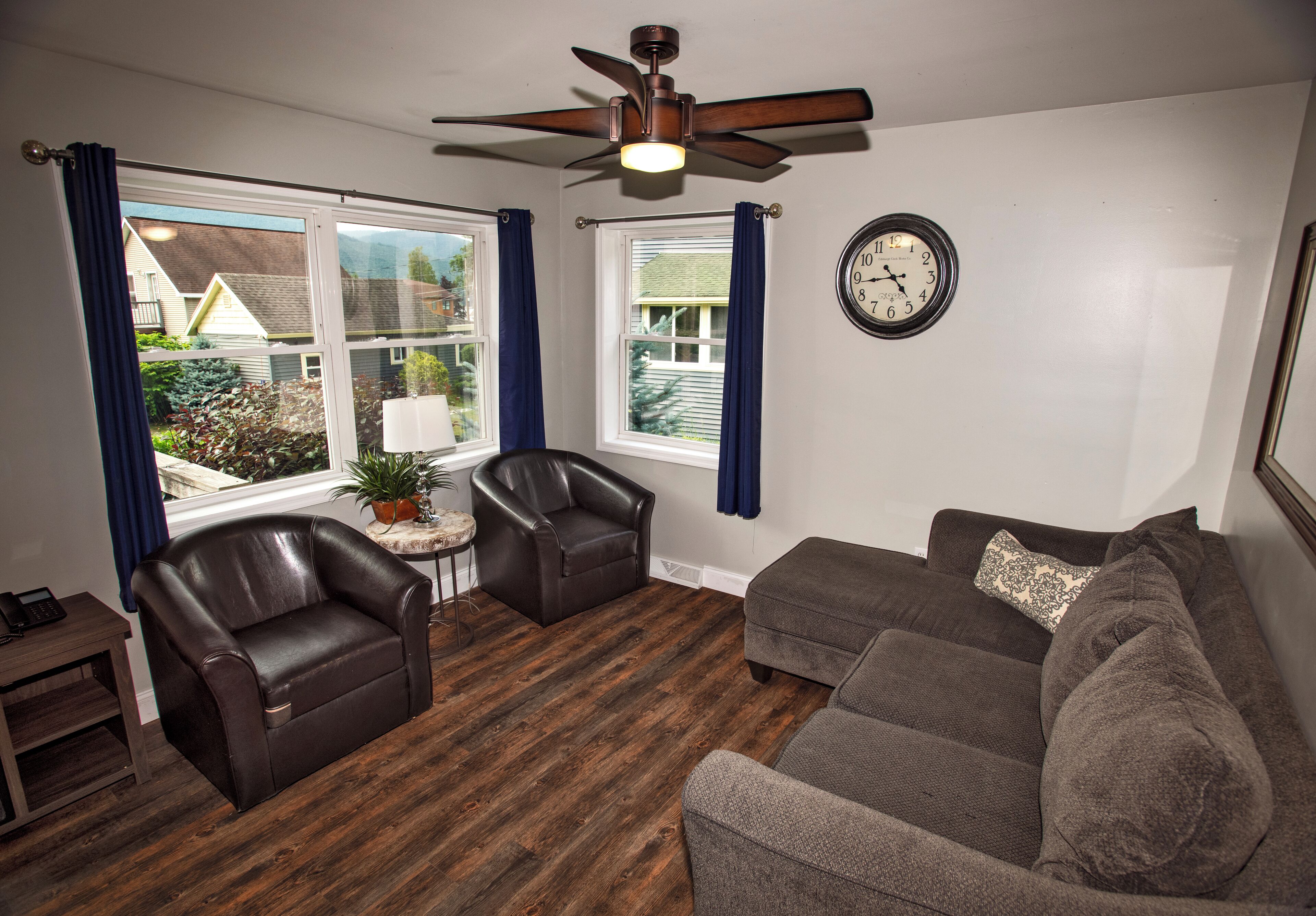 A living room with a brown leather chair, a gray sofa, a wooden floor, and a ceiling fan.