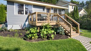A house with a wooden deck, a staircase, and a garden with various plants. - Beach Cottages at Lake George (Lake George)