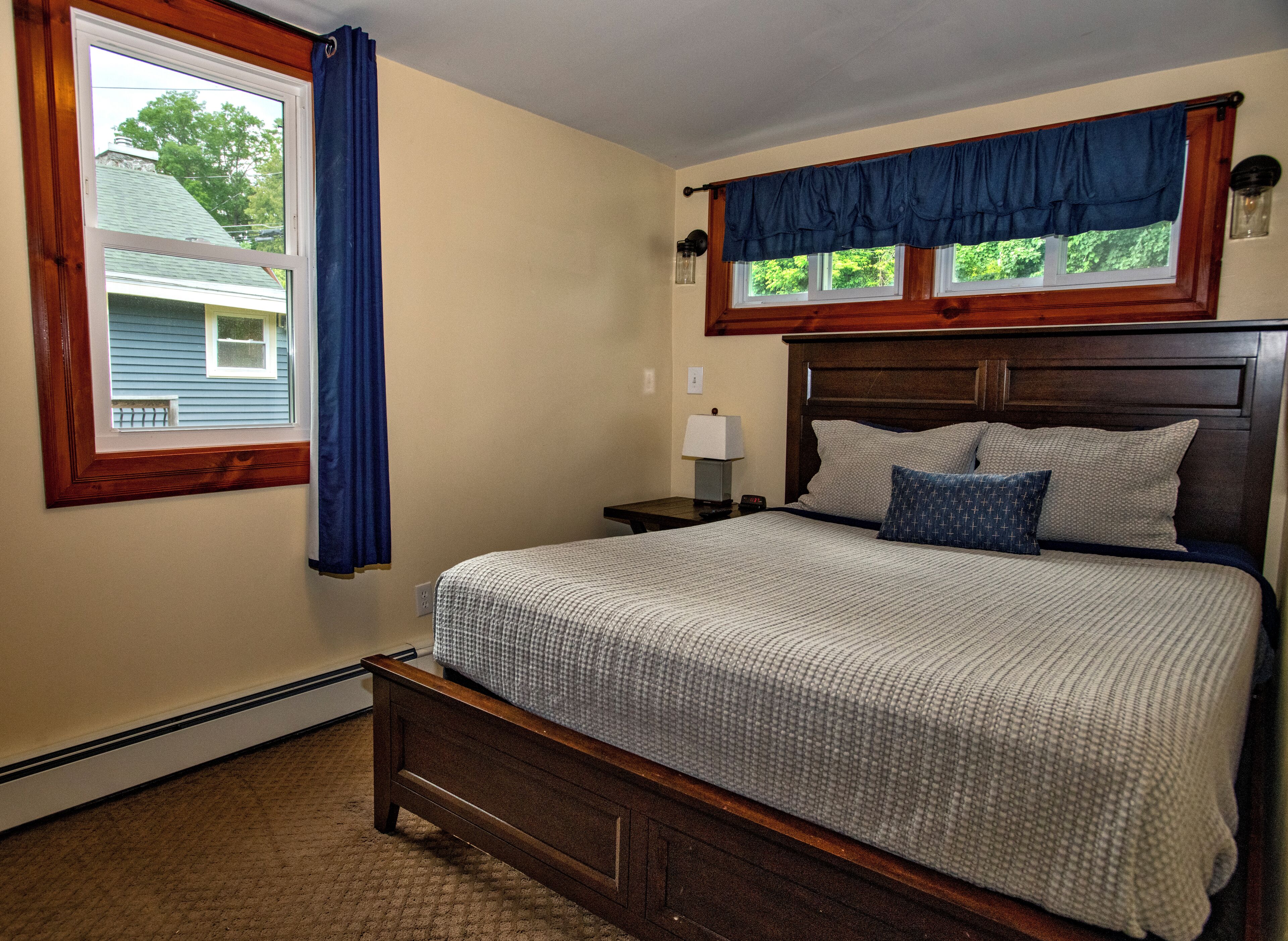 A bedroom with a wooden bed, a nightstand with a lamp, a window with blue curtains, and a view of a house outside.