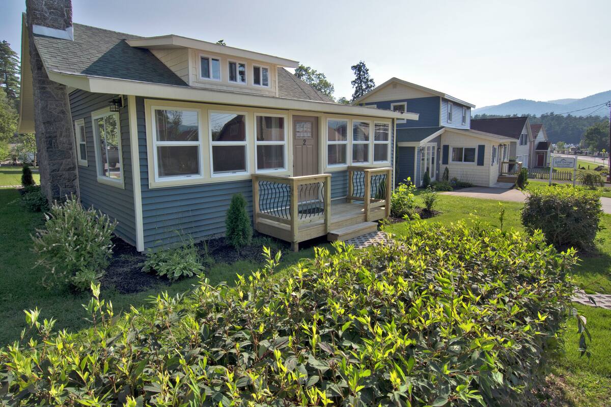 A two-story house with a covered porch and a small deck, surrounded by well-maintained gardens and a hilly landscape in the background.