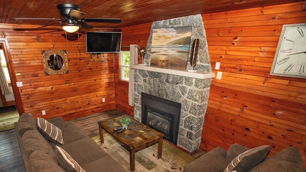 A cozy cabin interior with a stone fireplace, wooden walls, a ceiling fan, a flat-screen TV, and a large window. - Beach Cottages at Lake George (Lake George)