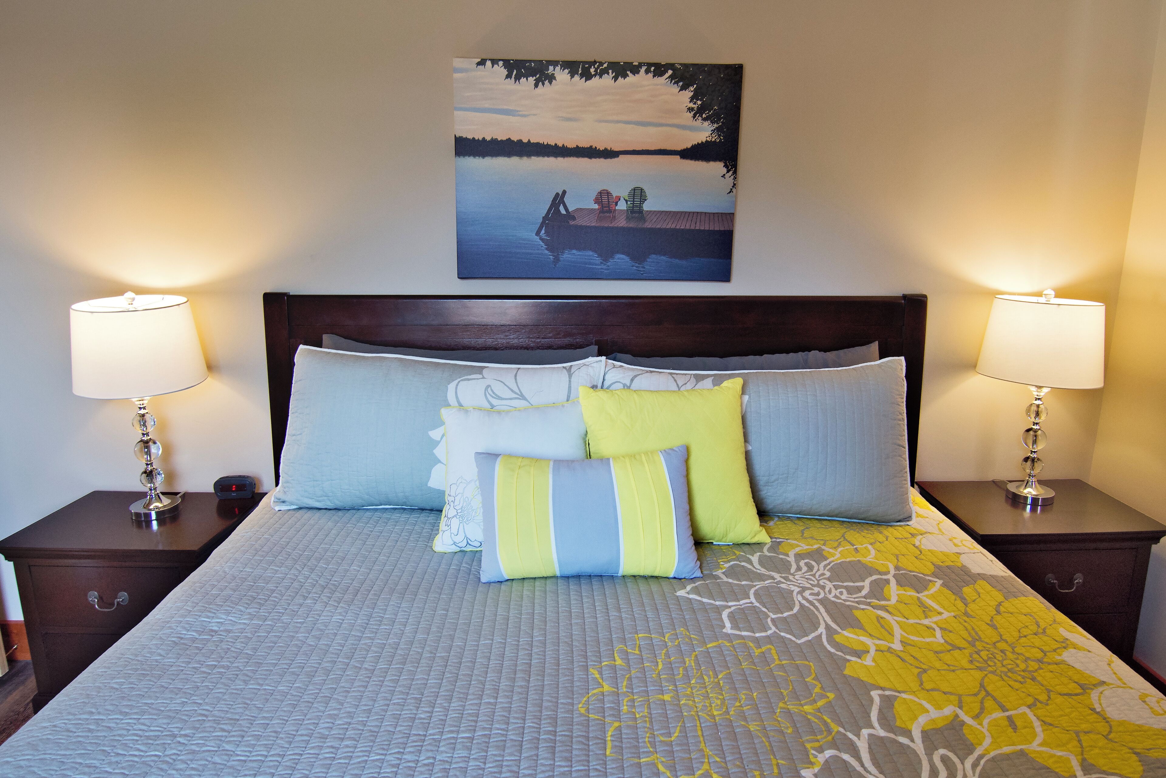 Un lit bien fait, recouvert d’une couvre-lit à motifs floraux, avec deux lampes de chevet et un tableau représentant un paysage lacustre au-dessus du lit.