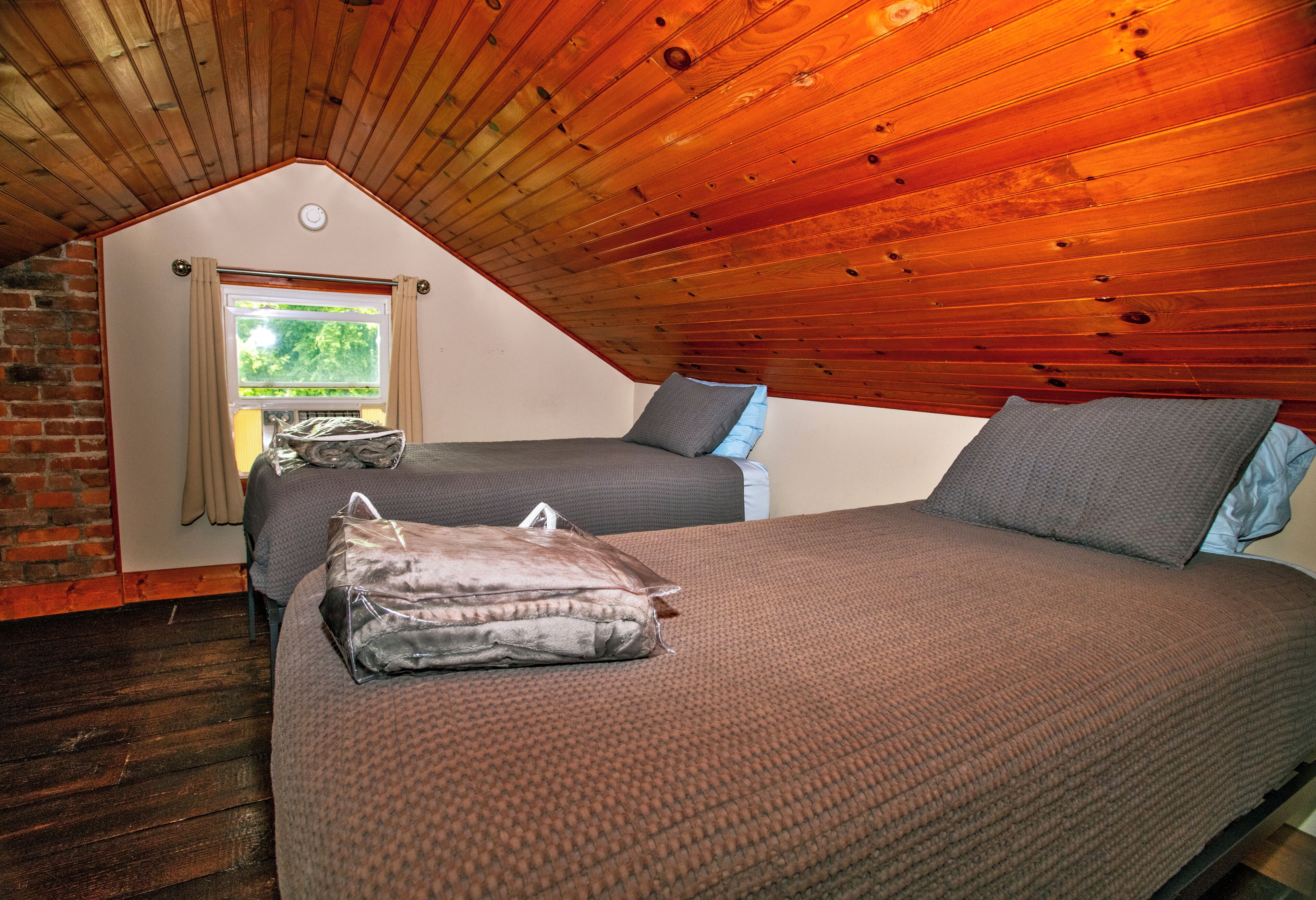 A cozy attic bedroom with a sloped wooden ceiling, two beds with grey bedding, and a window with curtains.