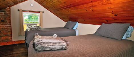 A cozy attic bedroom with a sloped wooden ceiling, two beds with grey bedding, and a window with curtains.