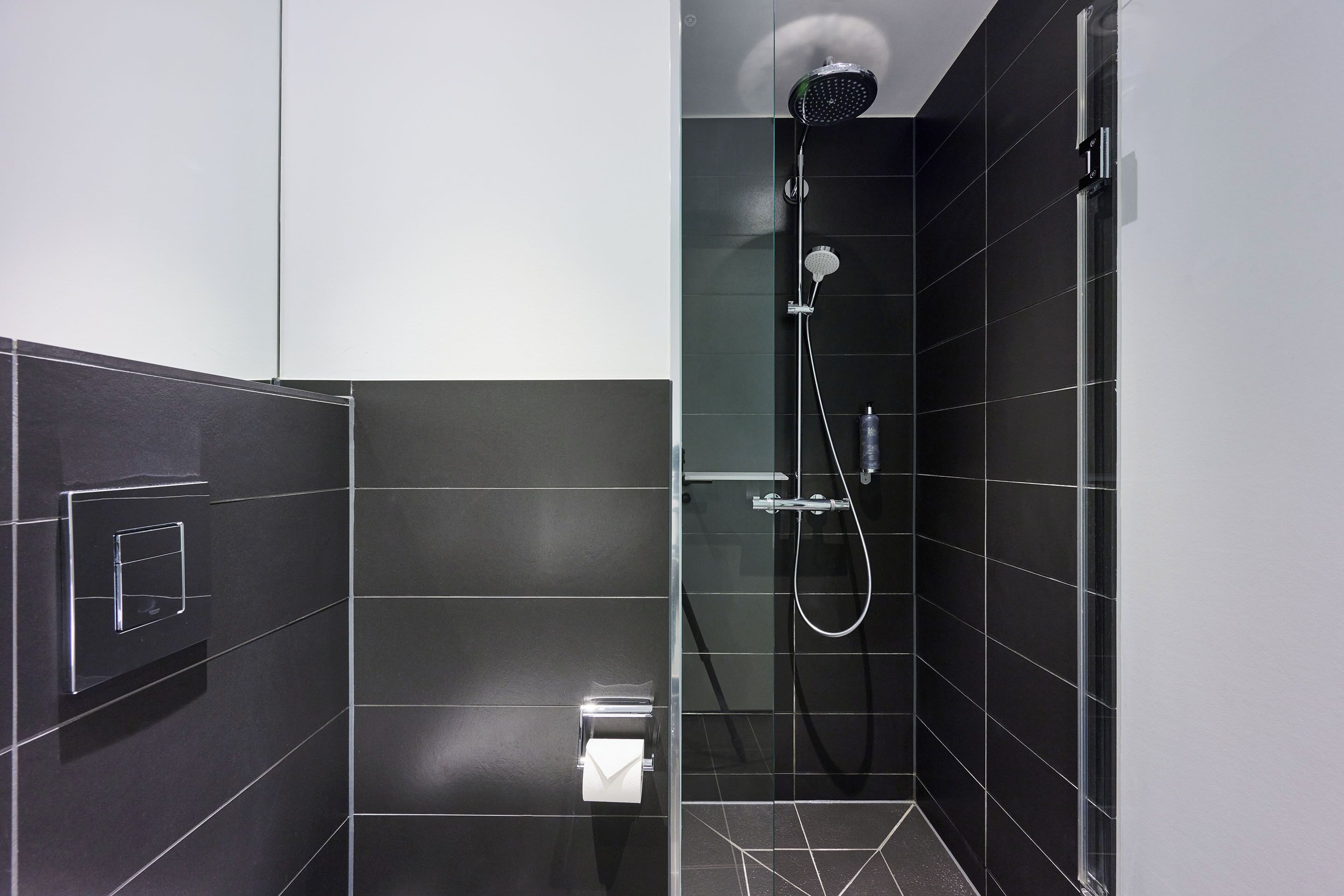 A modern bathroom with dark tiles, a glass shower enclosure, a handheld showerhead, and a towel dispenser.