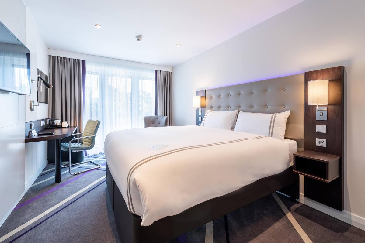 A hotel room with a large bed, a desk, a chair, a TV, and a window with curtains.