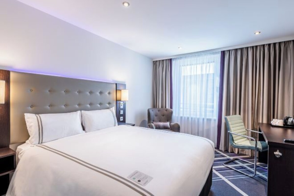A hotel room with a large bed, a headboard with lights, a desk, a chair, and a large window with curtains.