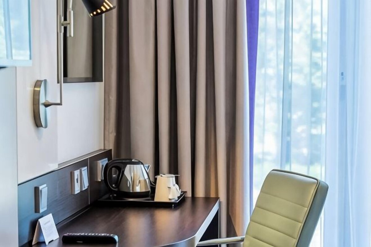 Ett hotellrum med ett skrivbord, en stol, en vattenkokare, ett bricka med en tekanna, en spegel och gardiner.