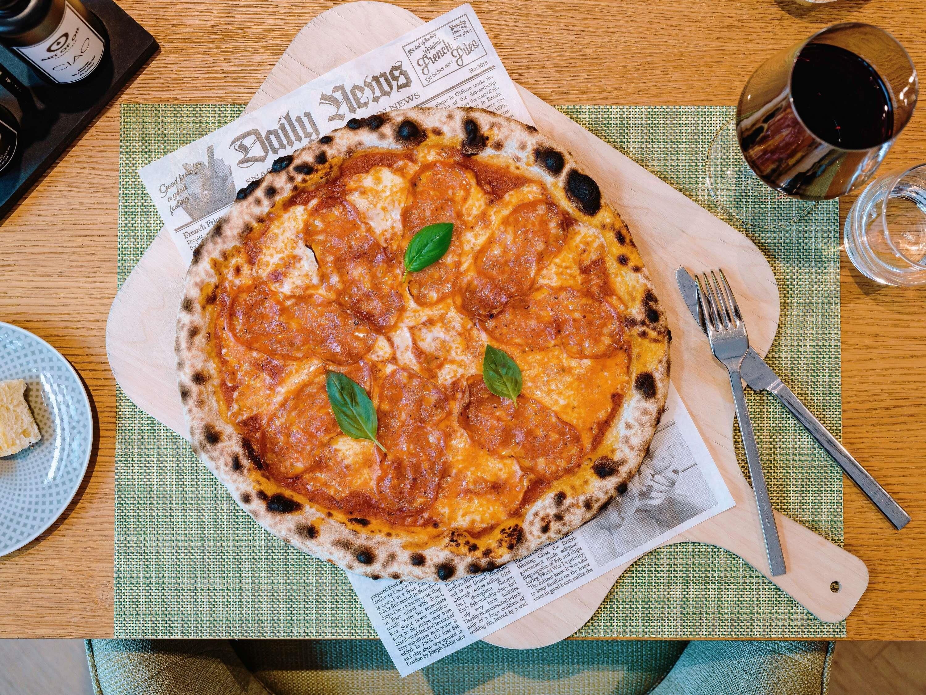 A pizza on a wooden board with a newspaper underneath, a fork, and a glass of red wine on the table.