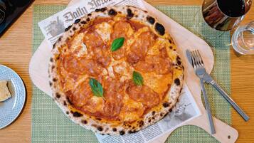 A pizza on a wooden board with a newspaper underneath, a fork, and a glass of red wine on the table.
