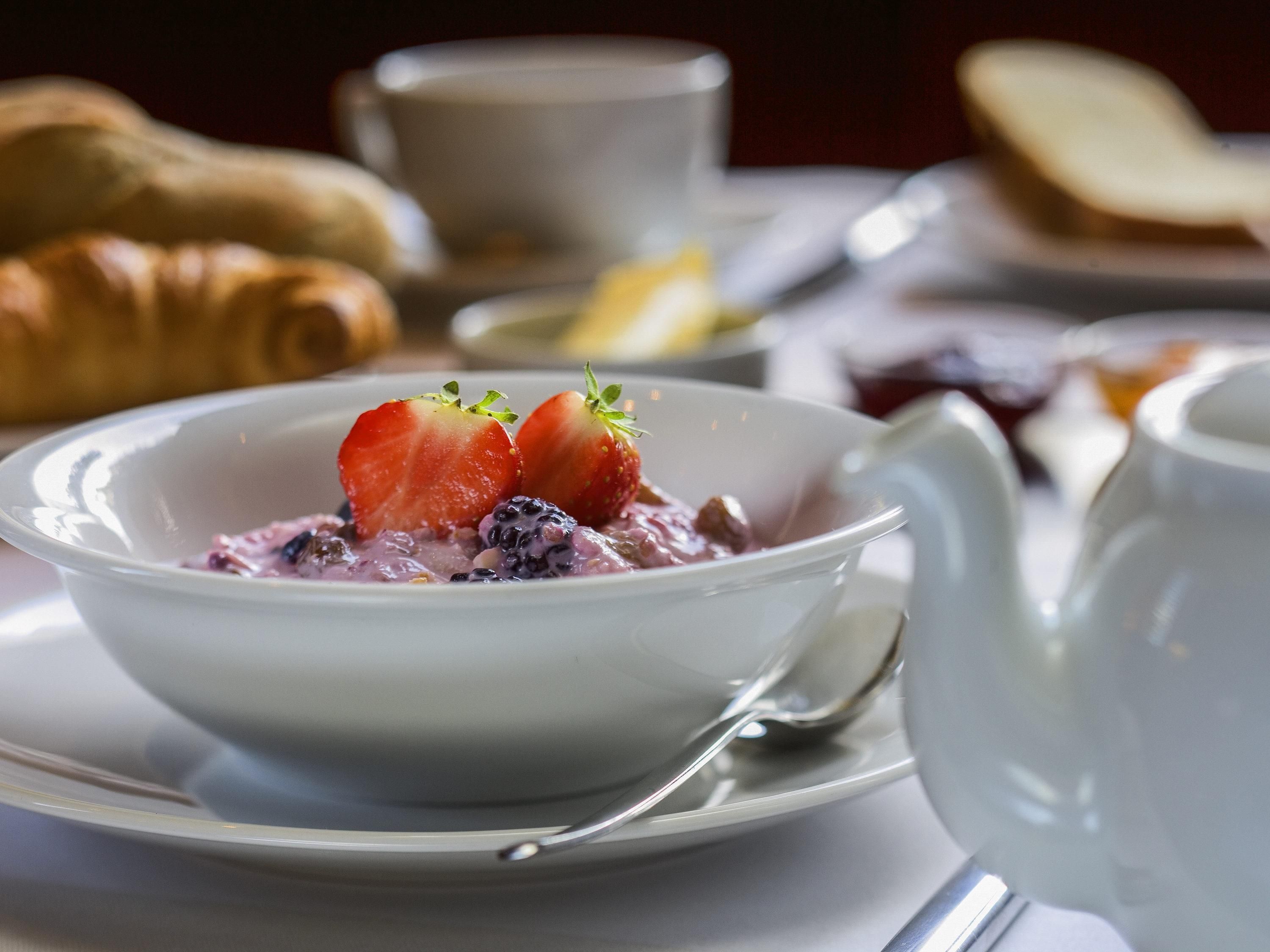 Una escena de desayuno con un tazón de avena cubierto de fresas y arándanos, un croissant y una taza de café sobre una mesa.