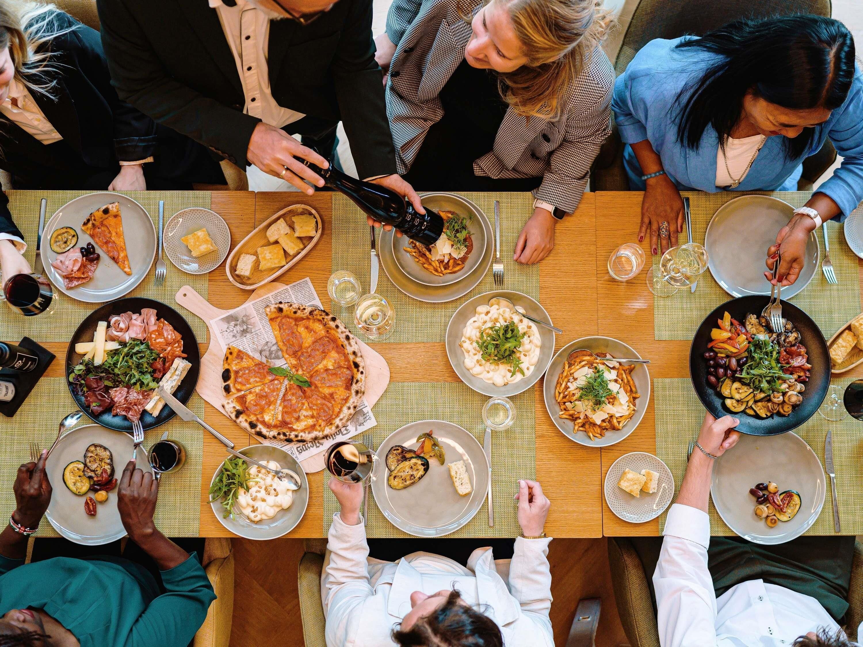 A group of people dining at a table with various dishes, including pizza, pasta, and salads.