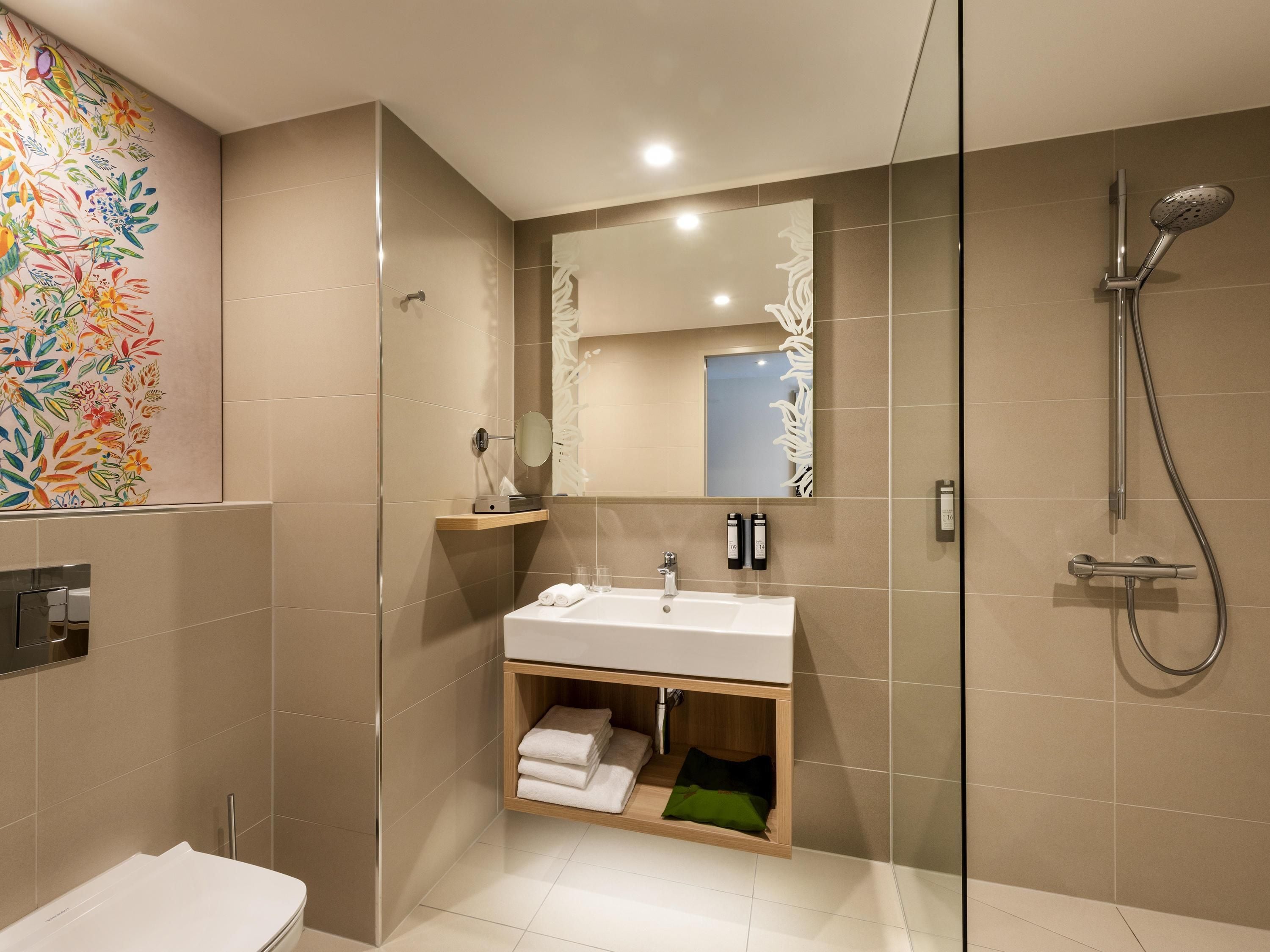 A modern bathroom with a glass shower enclosure, a sink with a mirror above, and a colorful floral wall mural.
