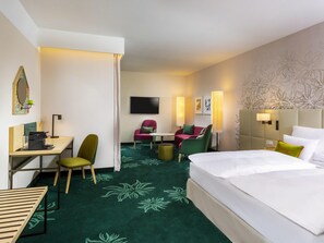 A hotel room with a bed, desk, chairs, a flat-screen TV, and a carpet with a floral pattern. - Mövenpick Hotel Stuttgart Messe & Congress (Leinfelden-Echterdingen)