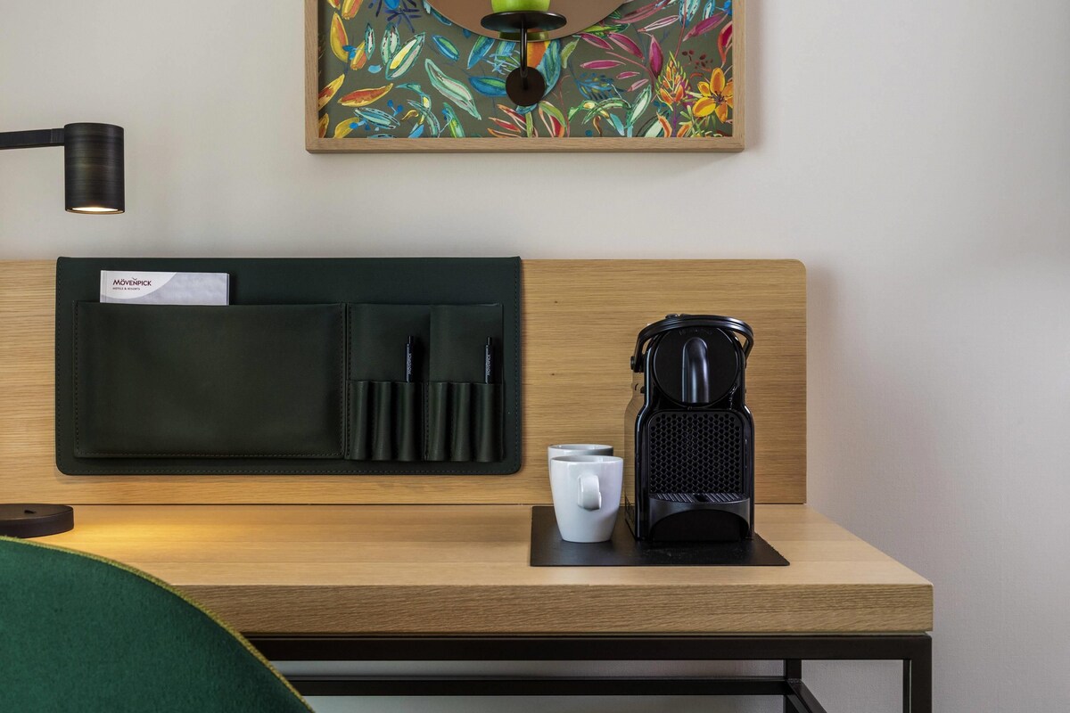 A neatly arranged hotel room with a wooden headboard, a black leather wallet with pens, a white mug, and a black coffee maker on a wooden table.