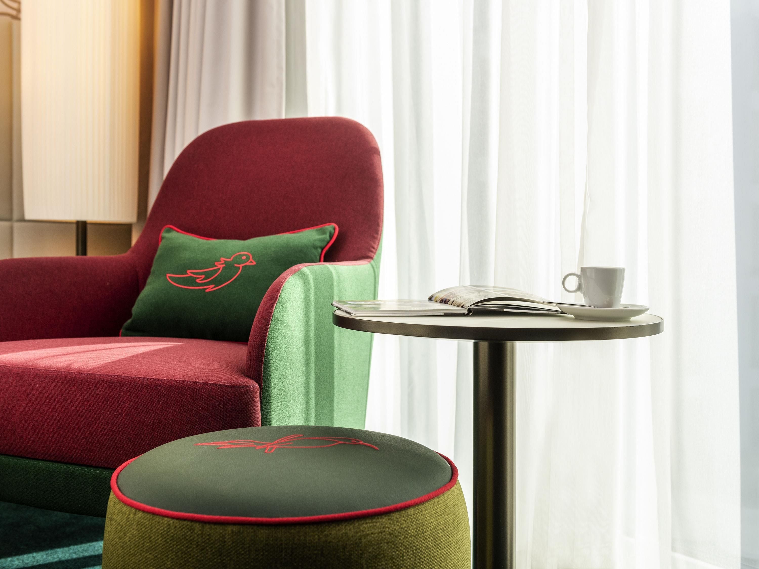 Un sillón rojo con un cojín verde y un otoman a juego, una taza blanca y un libro sobre una mesita redonda.