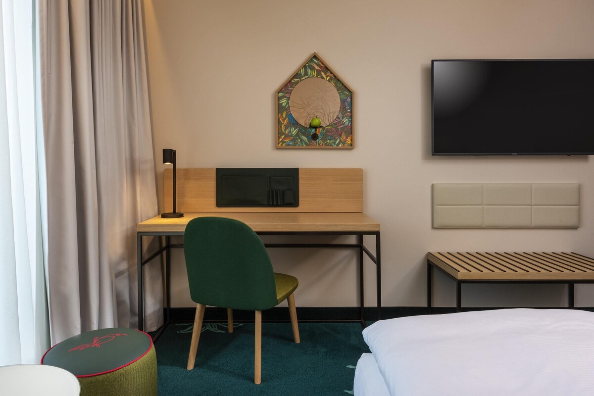 A hotel room with a bed, a desk with a chair, a television, and a wall-mounted artwork.