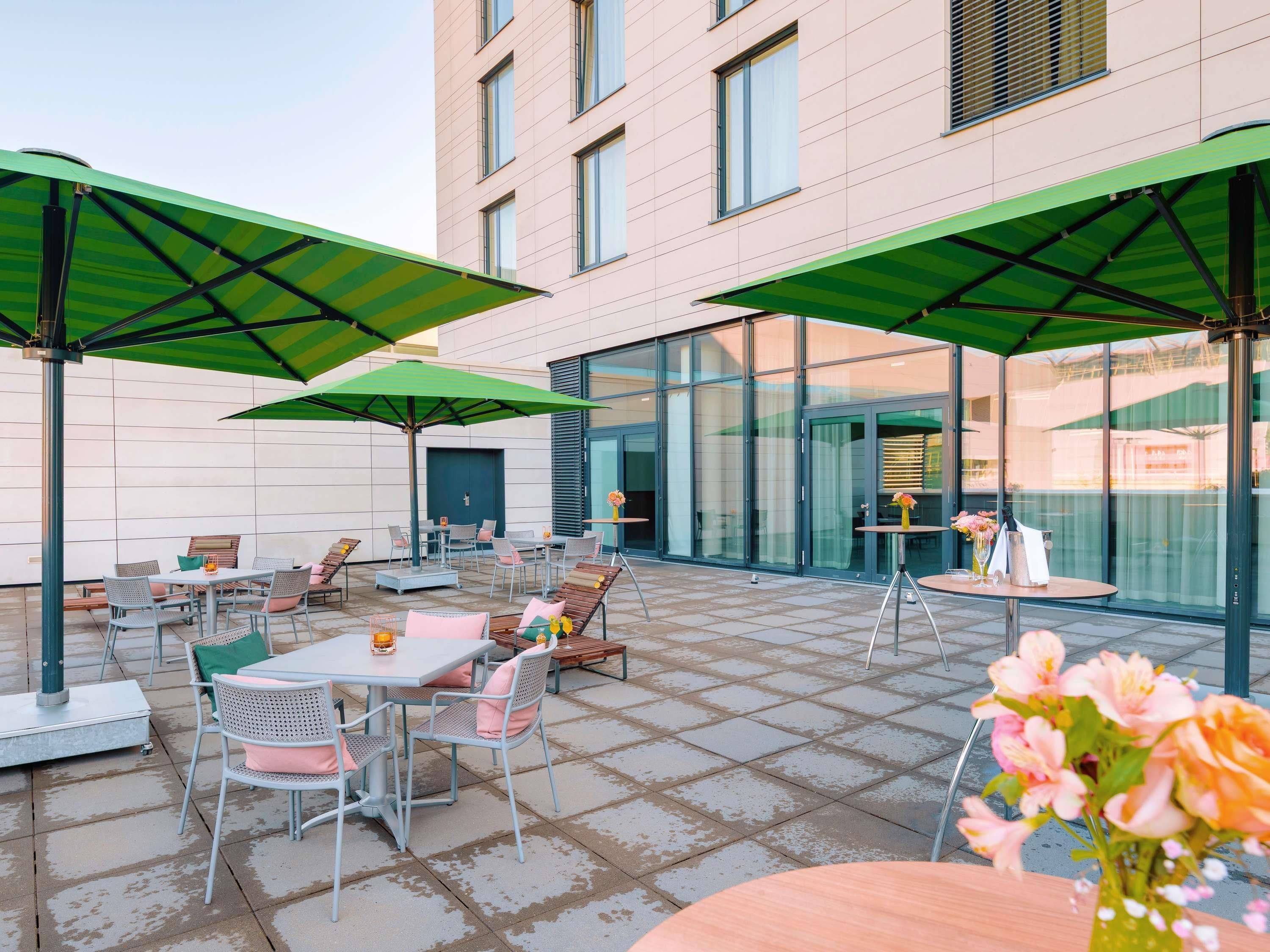 Eine Terrasse im Freien mit Tischen und Stühlen, grünen Sonnenschirmen und einem Gebäude mit großen Fenstern im Hintergrund.