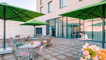 Eine Terrasse im Freien mit Tischen und Stühlen, grünen Sonnenschirmen und einem Gebäude mit großen Fenstern im Hintergrund.
