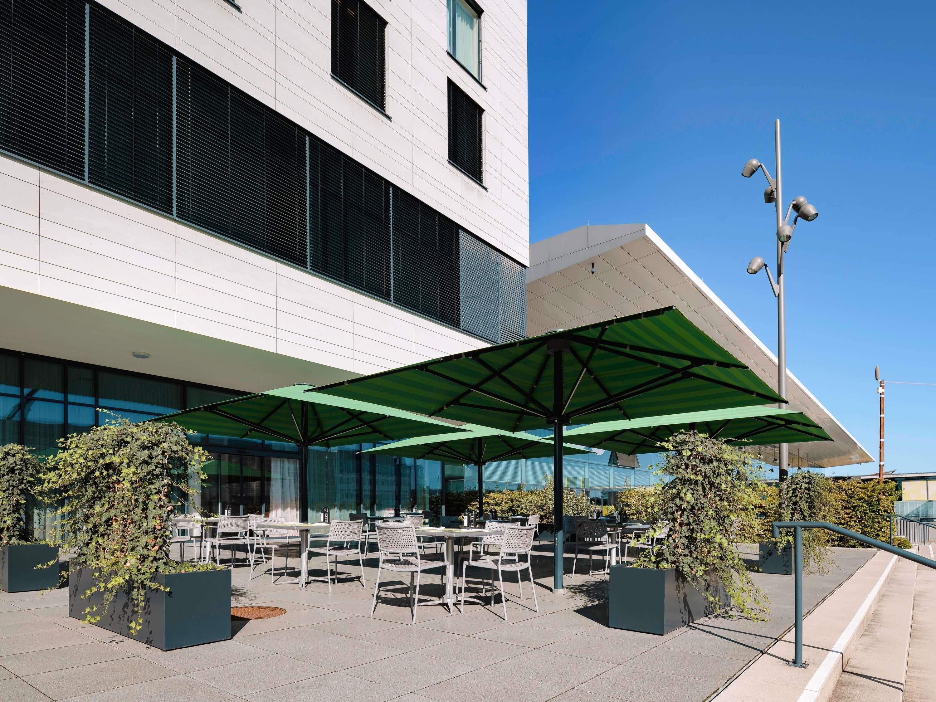 Un área de estar al aire libre con mesas y sillas, cubierta por sombrillas verdes, frente a un edificio moderno con grandes ventanales.