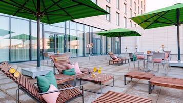 Un espacio al aire libre con mobiliario de madera, sombrillas verdes, ventanas amplias y un edificio moderno al fondo.