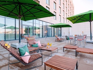 An outdoor seating area with wooden furniture and green umbrellas, large windows, and a modern building in the background. - Mövenpick Hotel Stuttgart Messe & Congress (Leinfelden-Echterdingen)