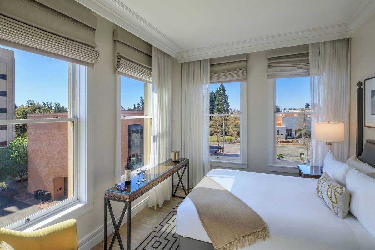 A hotel room with a large bed, a desk with a lamp, a chair, and a view of the city.