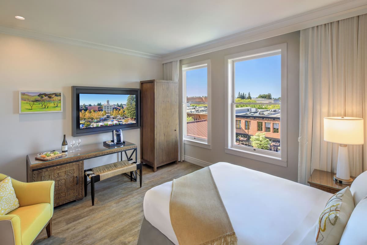 A hotel room with a bed, a desk with a television, a chair, and large windows with a city view.