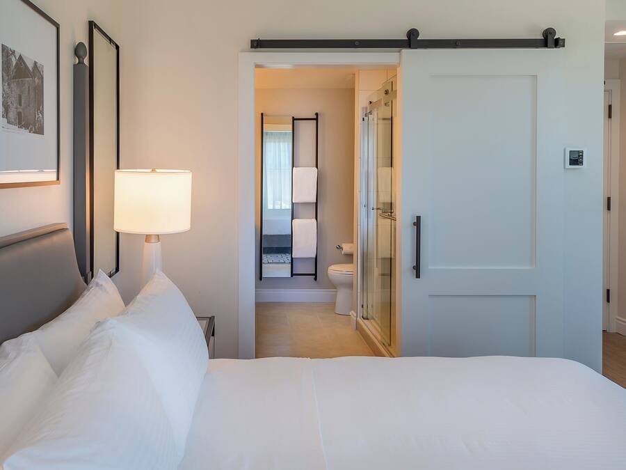 Et hotelværelse med en stor seng, en sengebordlampe, et billede i ramme på væggen og en skydedør til et badeværelse.