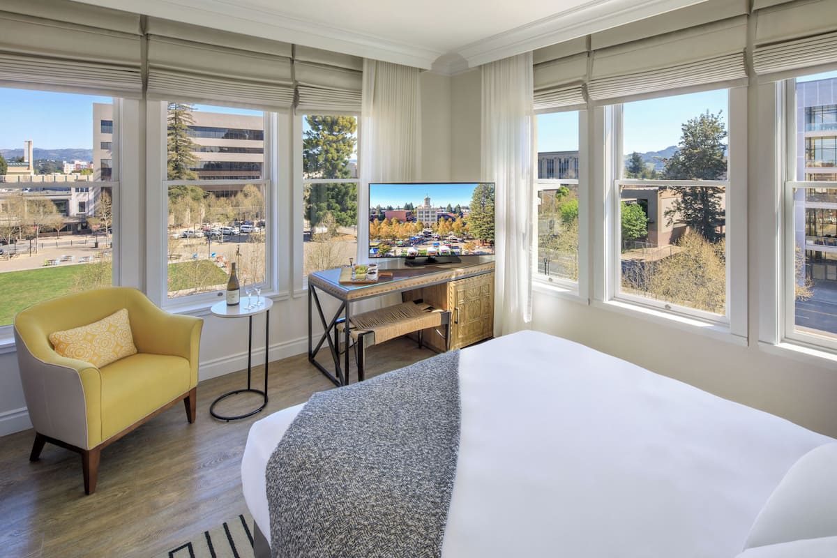 A hotel room with a bed, a desk, a chair, a TV, and large windows offering a city view.