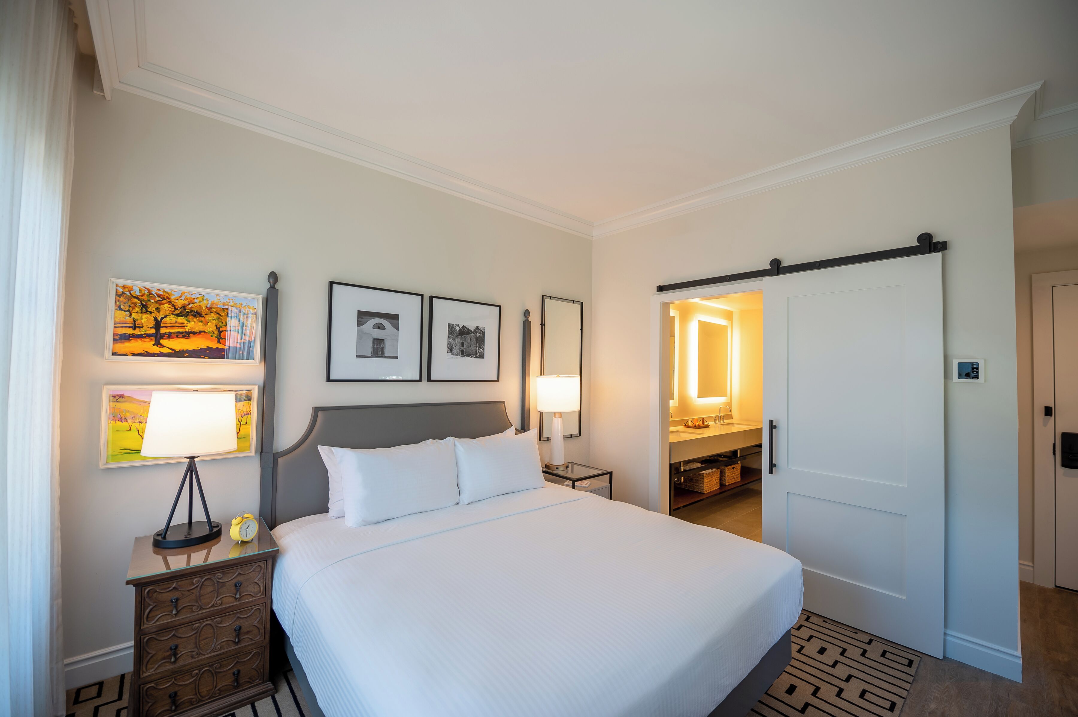 A hotel room with a bed, a nightstand, a lamp, a dresser, a mirror, and a door leading to a bathroom.