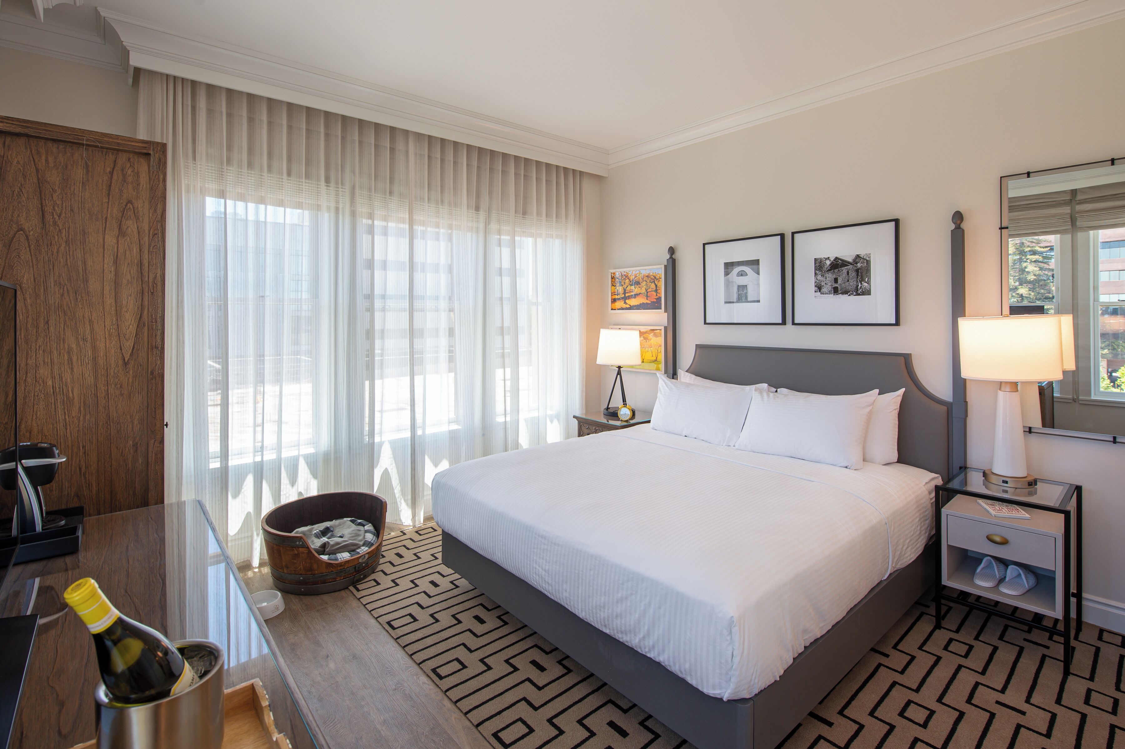 A hotel room with a large bed, a wooden desk with a bottle of champagne, a small bucket, and a nightstand with a lamp.