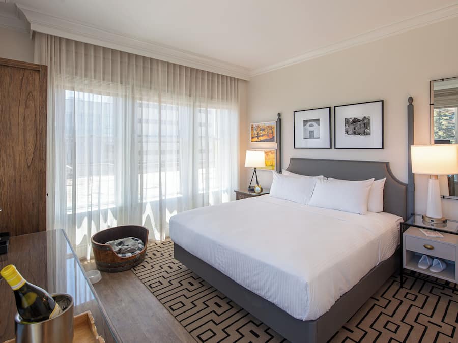 A hotel room with a large bed, a wooden desk with a bottle of champagne, a small bucket, and a nightstand with a lamp.